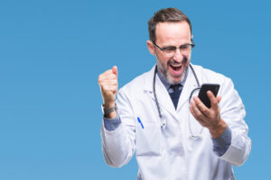 Middle age senior hoary doctor man texting using smartphone over isolated background screaming proud and celebrating victory and success very excited, cheering emotion