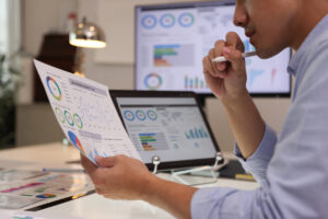 Focused businessman analyzing marketing reports on a desktop monitor and reviewing paper graphs, financial stats, and startup project infographics.